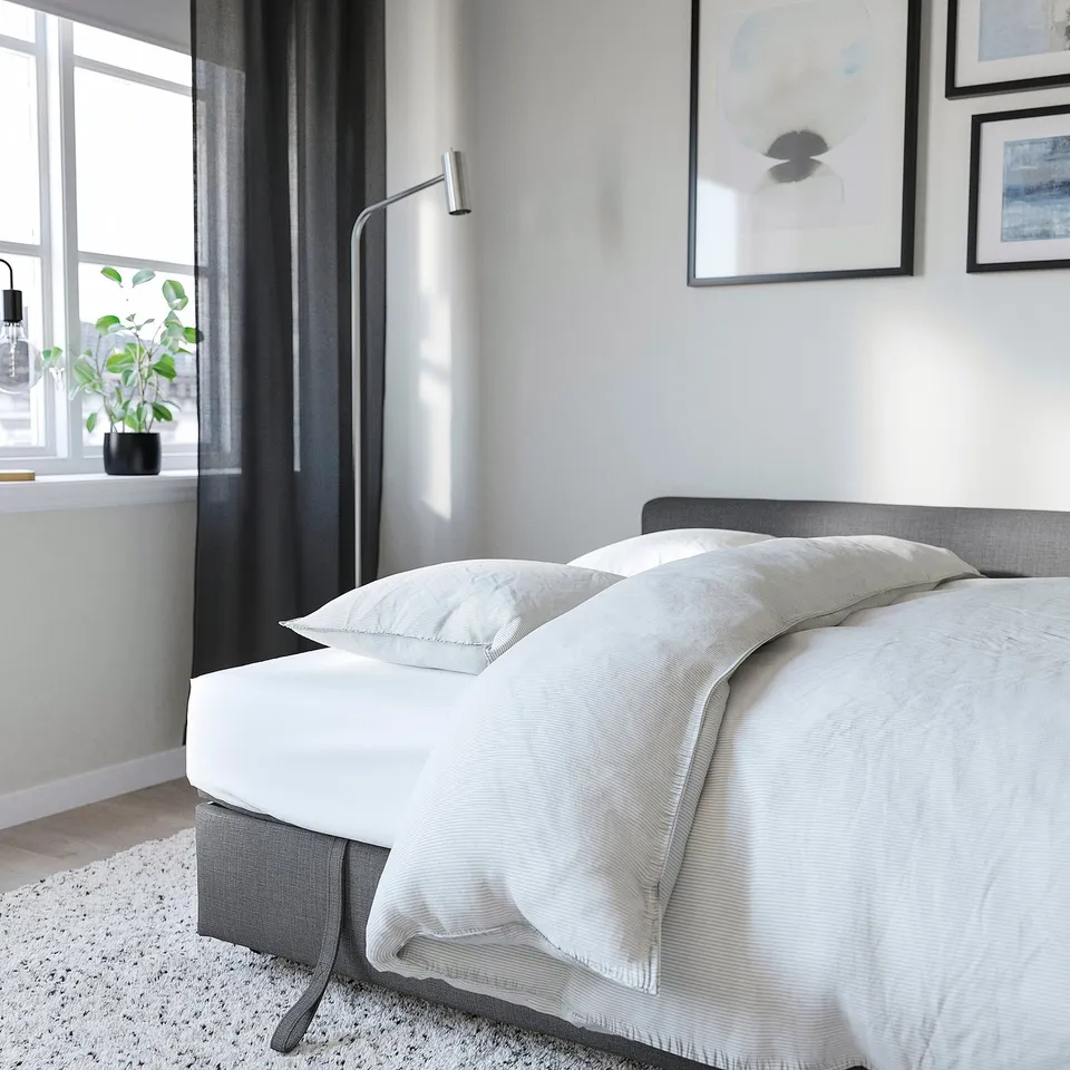 A modern bed with white linens, grey headboard, and wall lamp, offering storage and sofa conversion.