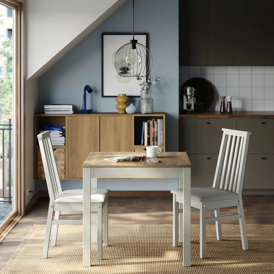 ÅLHULT dining set with light grey chairs and white table, beside wooden cabinet with books and kitchen counter coffee maker.