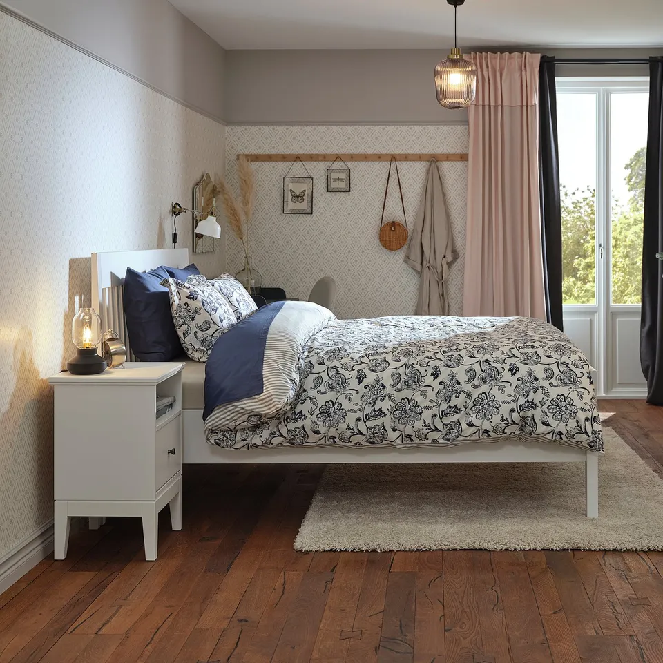 Modern bedroom with IDANÄS white bed, blue comforter, floral sheets, wooden floor, and pink curtains.