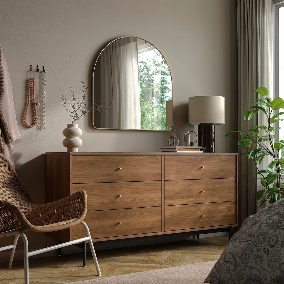 Walnut chest with 6 drawers, brass knobs, black legs, mirror, lamp, wicker chair.