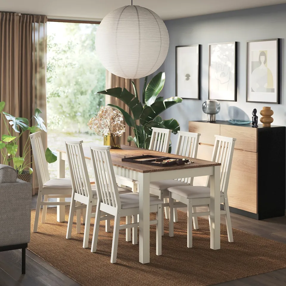 Modern dining area with ÅLHULT white chairs, cushioned seats, pine frame, table, window, plants, artwork, sideboard.