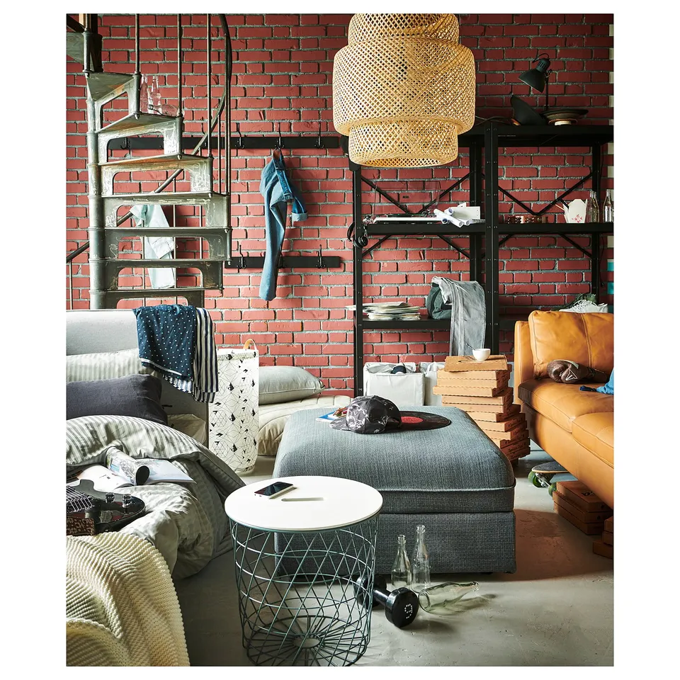 Cosy living room with brick wall, spiral stairs, grey sofa, & black metal shelves with various items; hanging lamp.