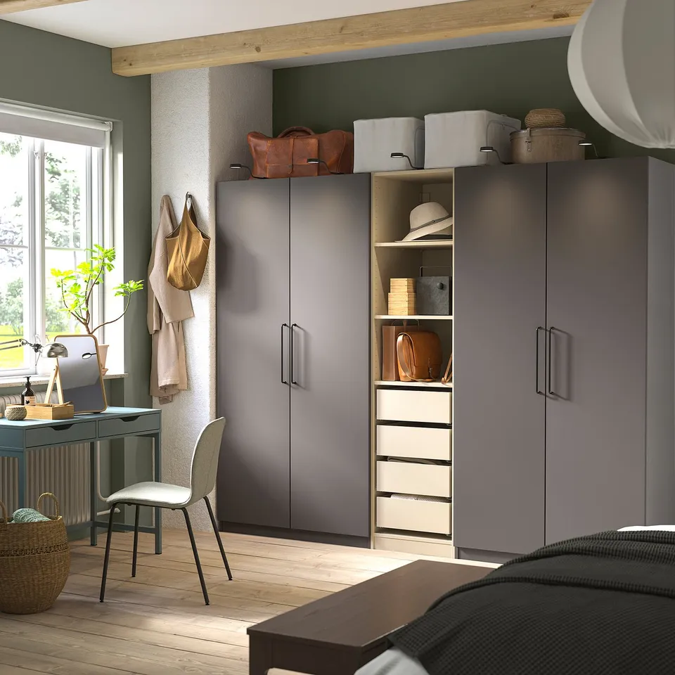 Grey PAX wardrobes by green wall, sleek handles, white shelving. Small desk, chair, plant, window light. Wooden floors, exposed beams.