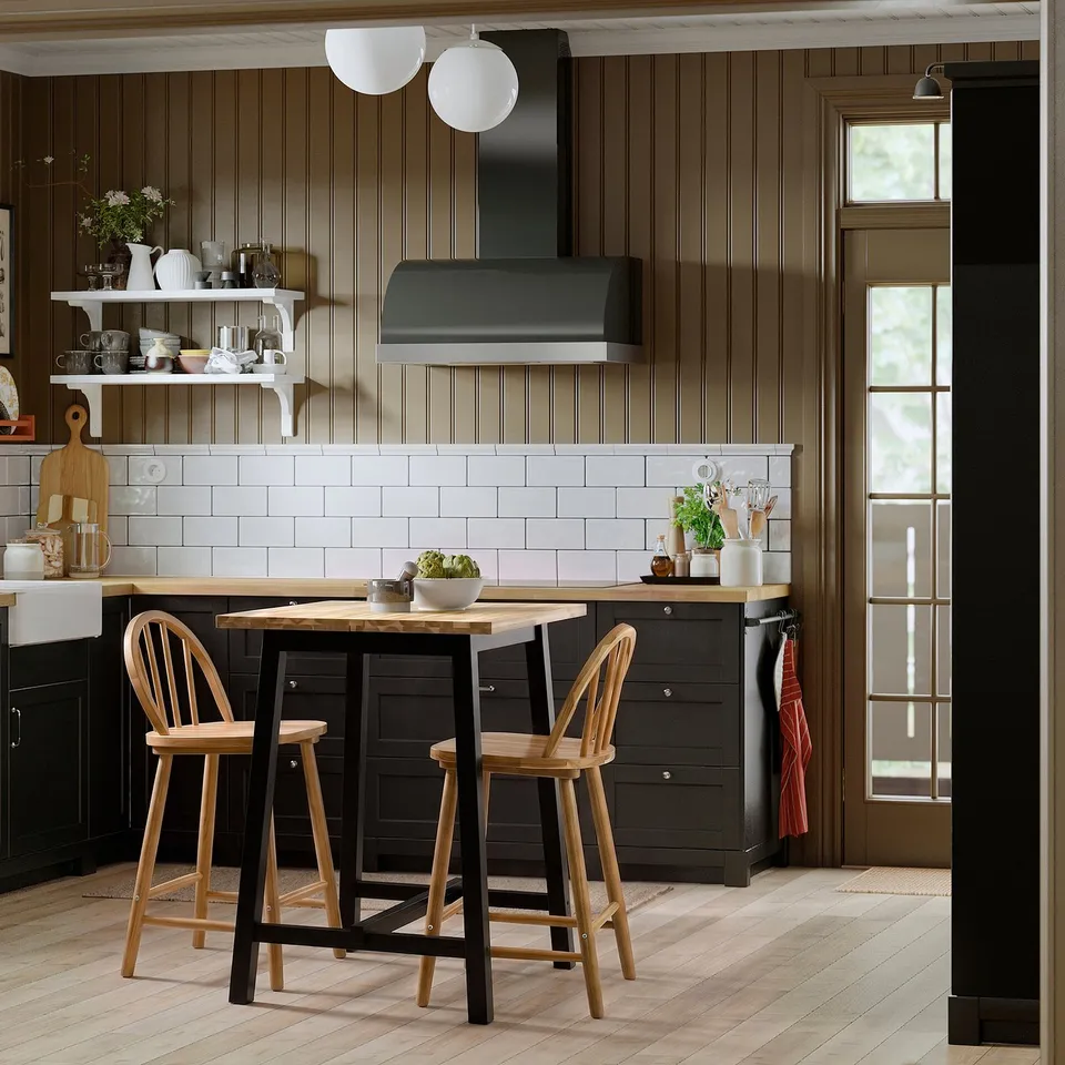 Modern kitchen with SKOGSTA table and stools, dark cabinets, white countertops, and open shelving with dishware and flowers.