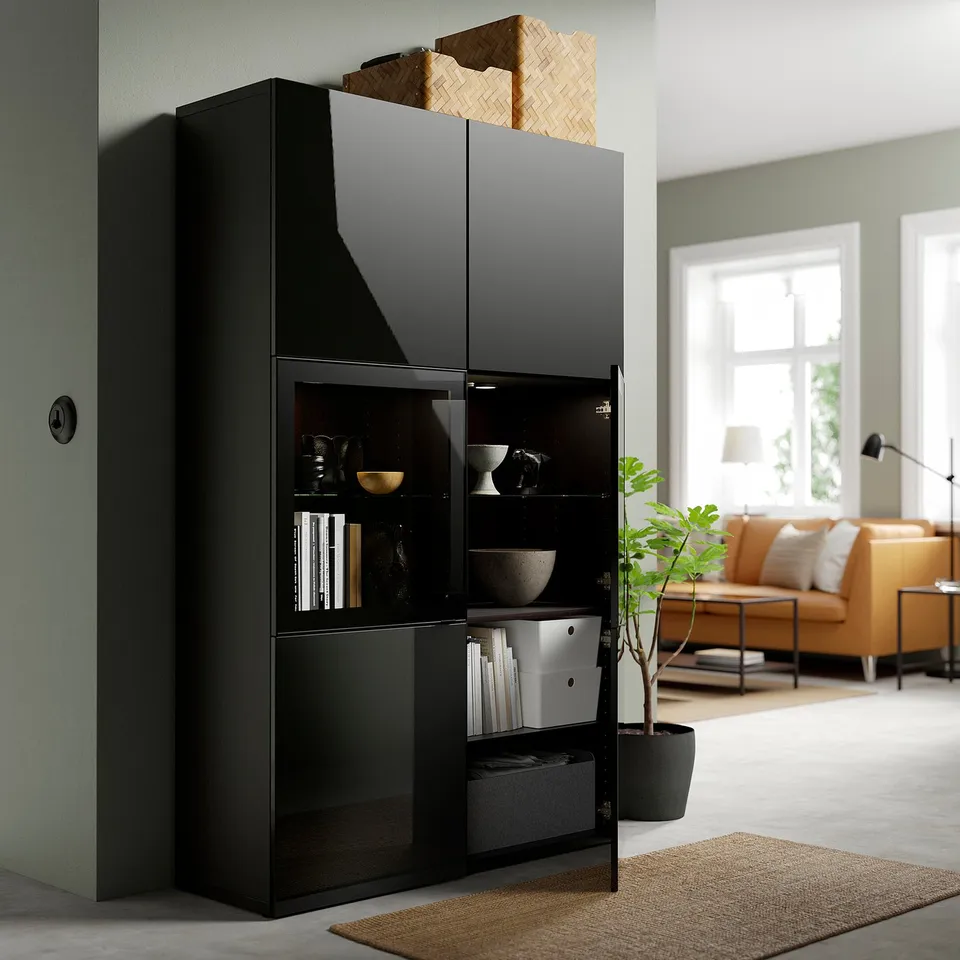 Black BESTÅ storage unit with glass doors, displaying books and decor, adjacent to a cosy living room with an orange sofa and a large window.