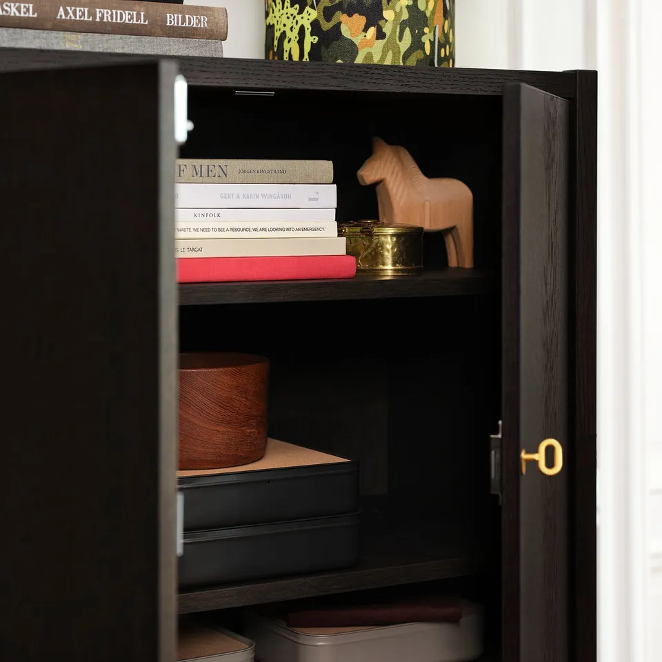 A dark wood cabinet with two adjustable shelves, holding books and decorative items, and featuring soft-close doors with a brass keyhole lock.
