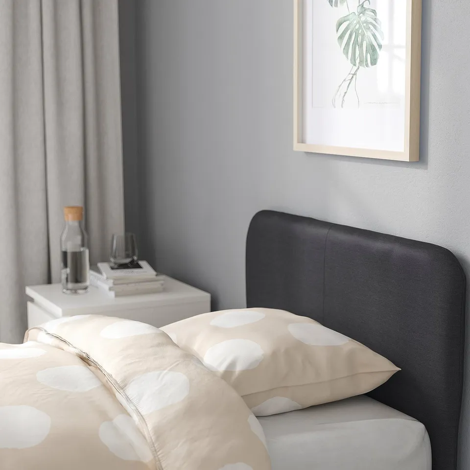 Bedroom scene with modern bed, black headboard, white polka dot bedding, grey wall, framed plant art, white bedside table, books, carafe, and glass.