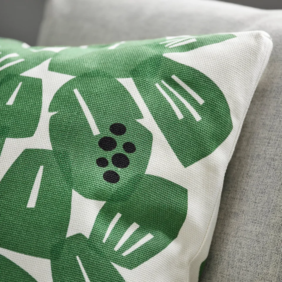 Close-up of a square throw pillow with vibrant green leaf patterns on white fabric, showcasing its thick, textured material and neat boxed corners.