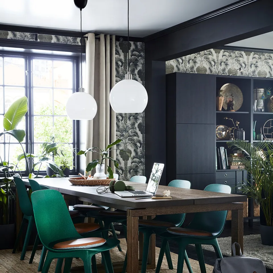 Modern dining area with wooden table, green chairs, and two white globe pendant lamps. Dark walls, patterned curtains, and plants.