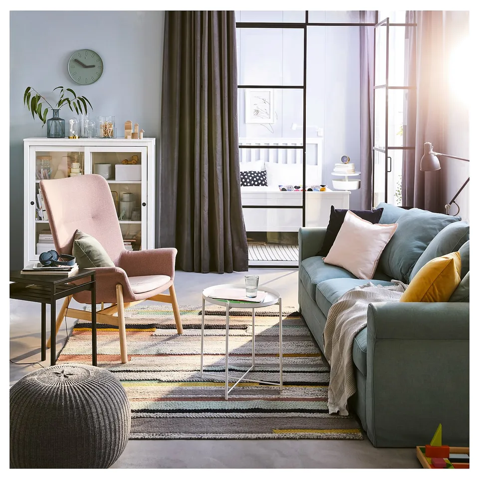 Bright living room with modern furniture: grey sofa, pink armchair, glass side table, colourful rug, and a curio cabinet.