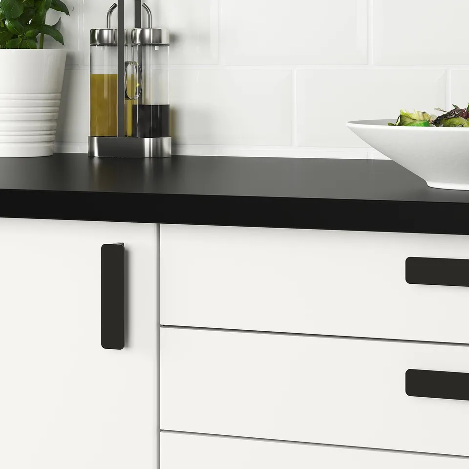 Modern kitchen with sleek black countertop and white cabinets accented with black handles, featuring a bowl of salad.