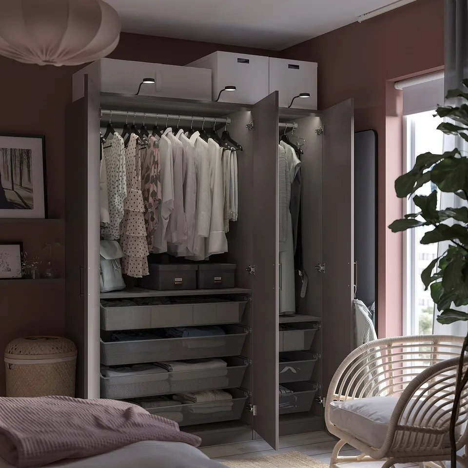 Modern bedroom with open dark grey PAX wardrobe displaying clothes, storage bins, and mesh drawers. Includes a wicker chair and large window.