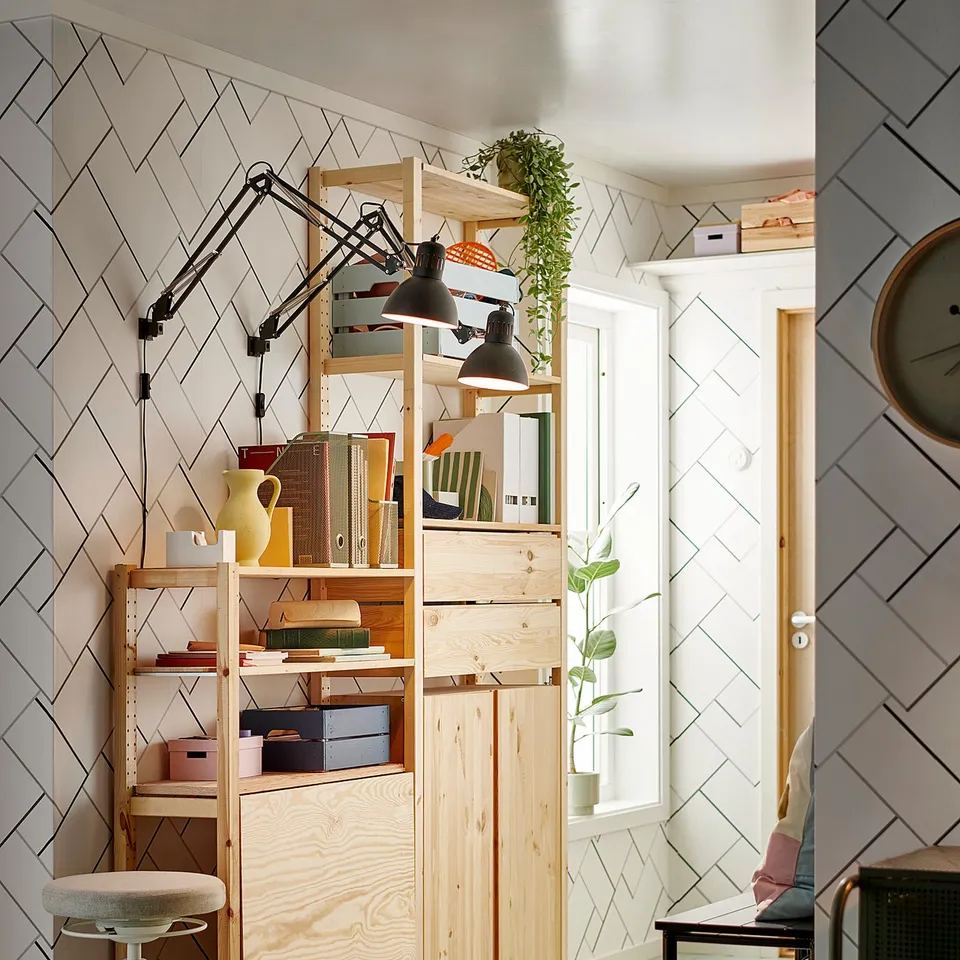 A modern office with an IVAR pine storage unit, white stool, adjustable lamps, plants, and a small black rack.
