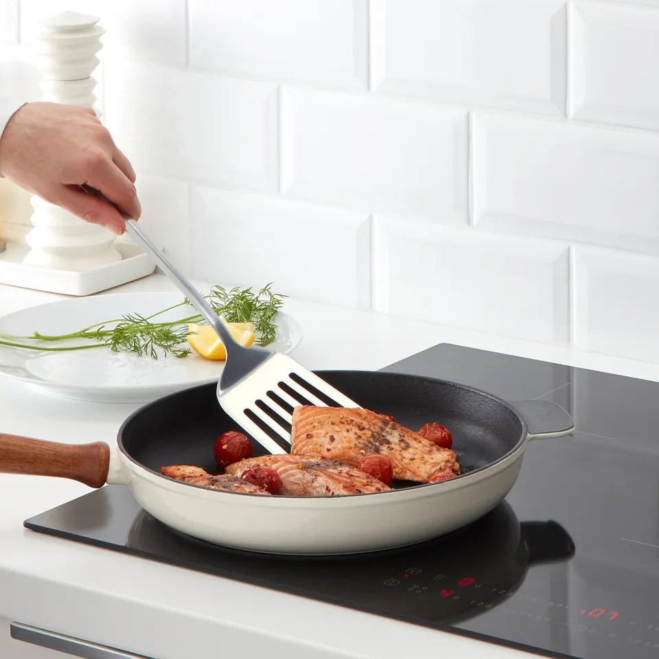 Person cooking fish with tomatoes in white frying pan on stove.