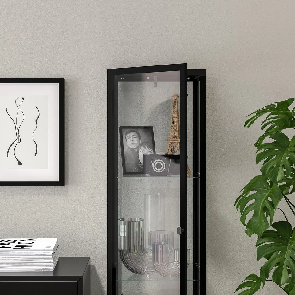 Black glass-door cabinet displaying decor items, with a light inside, positioned against a grey wall.