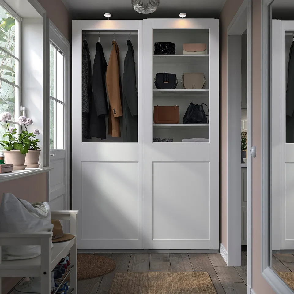 A modern white PAX wardrobe, tall with glass doors. It holds coats and handbags neatly. A chair sits nearby, small in comparison.