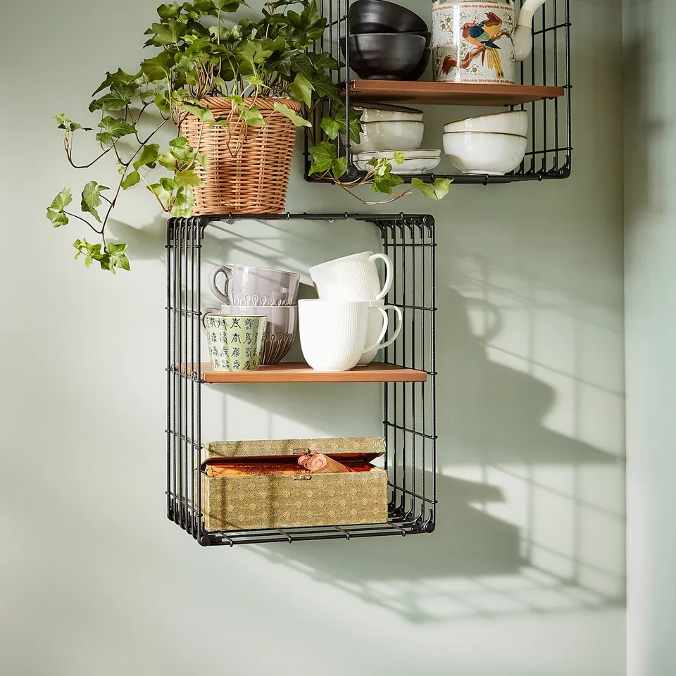 A wall shelf, GULLHULT in black metal and wooden shelves. Dishes, mugs, and plants against a green wall.