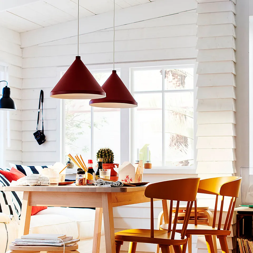 Bright living space with wooden table, chairs, and red pendant lamps. Large windows let in sunlight, light-colored rug on floor.