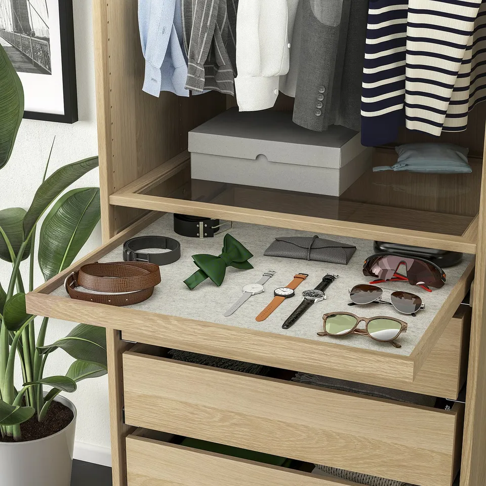 Wooden closet with sliding tray showcasing belts, watches, and sunglasses on a grey drawer mat, with hanging clothes and a plant beside.