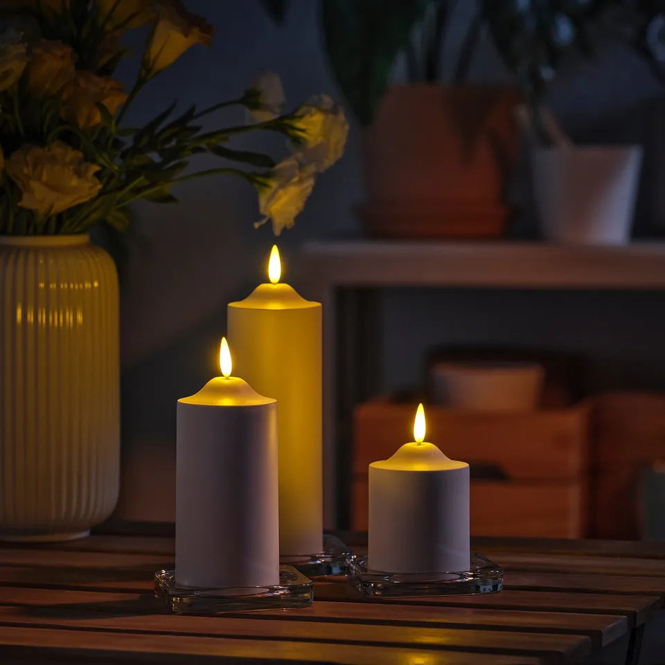 Three SMÅLOM led candles of varying sizes on table, with vase of flowers.
