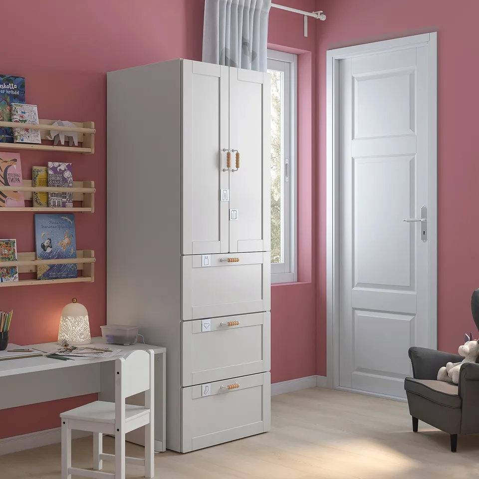 Kids room with pink walls, white wardrobe, shelves, desk, chair, lamp, and armchair.