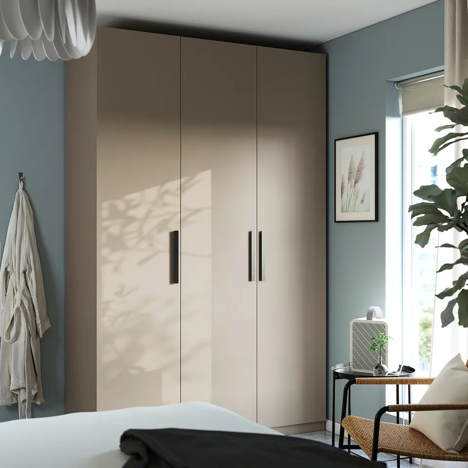 Modern bedroom with beige PAX wardrobe, blue walls. Wardrobe has black handles, left hooks, right plant, lamp, picture.