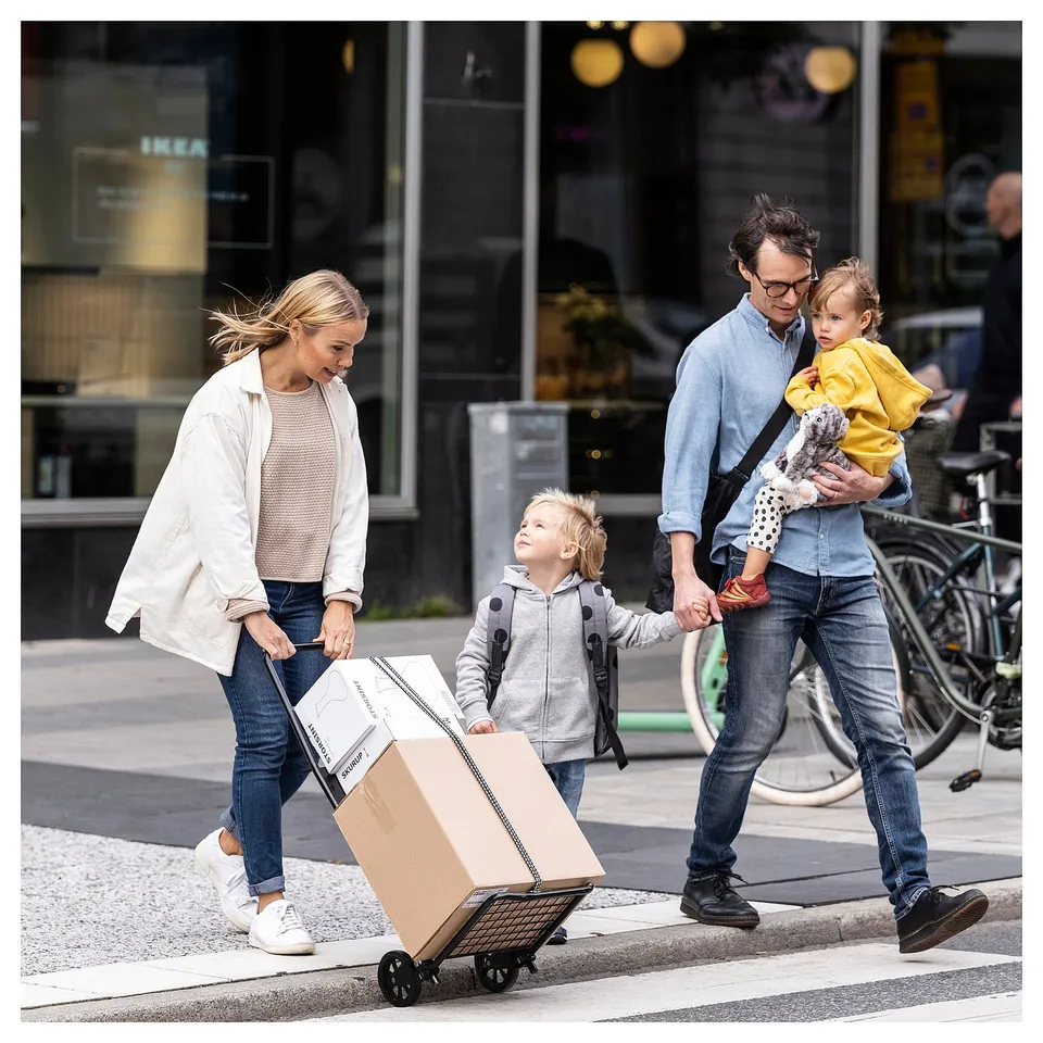 A family crosses a street with shopping from IKEA, using a beige RULLEBÖR trolley, with cardboard boxes, and two children.