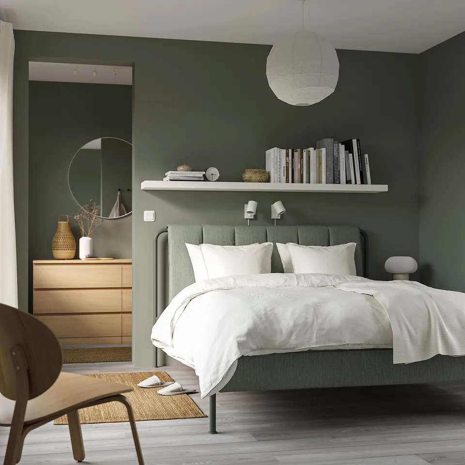 Modern bedroom with TÄLLÅSEN gray-green bed, white linens, dark green walls, wooden dresser, mirror, chair, rug, and hanging light.
