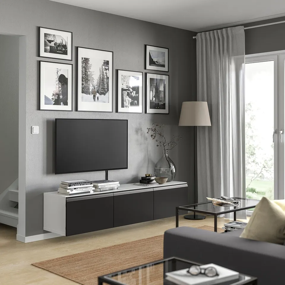 Modern living room with BESTÅ tv bench in black and white, featuring a flat screen tv, framed photos, grey curtains, and a glass coffee table.