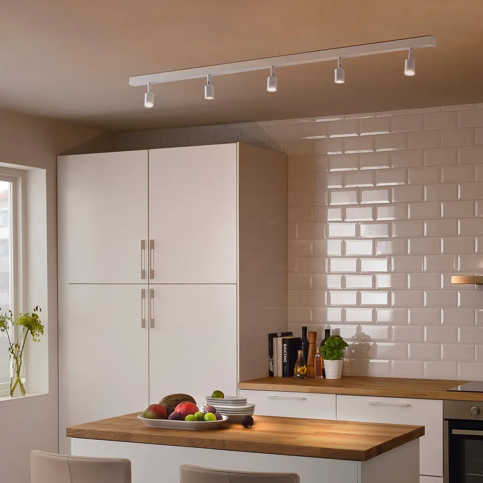 Modern kitchen with BÄVE ceiling light, wooden counters, and sleek cabinets. Fruits, plant, and décor complete minimalist style.