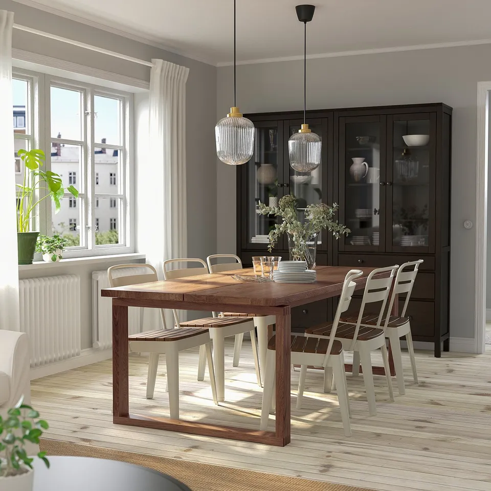 Modern dining set with large oak table, eight chairs, pendant lamps, and wooden cabinet with dinnerware. Bright room with large windows.