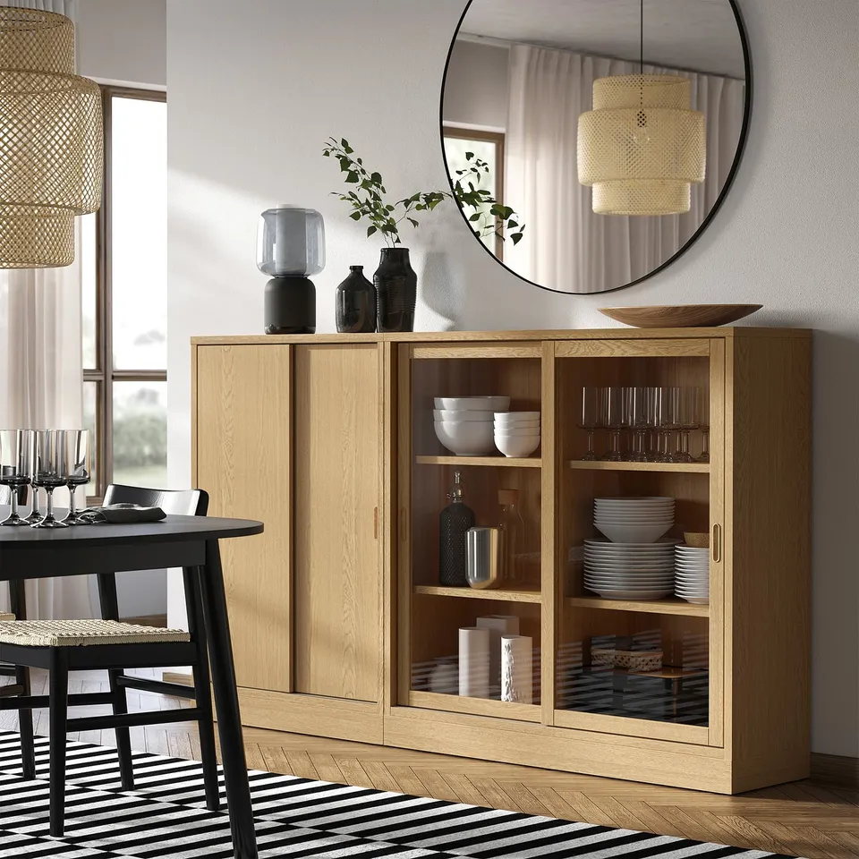 Wooden cabinet with glass doors showing dishes & vases. Right of dining table with wine glasses & napkin. Bright room with natural light.