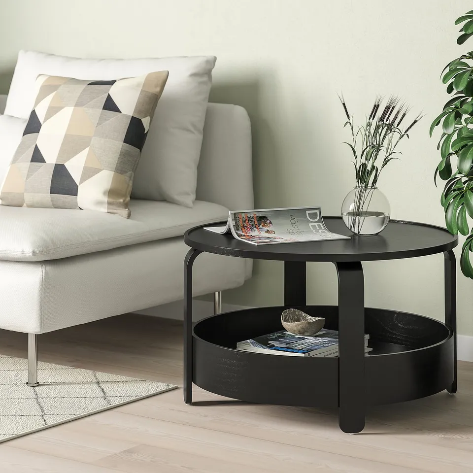 Modern black wooden BORGEBY coffee table next to large sofa. Circular shape with storage shelf and dried plant decoration.