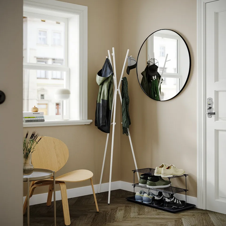 Entryway scene with wooden chair, white shoe rack holding multiple shoes, black coat stand holding jackets, and circular wall mirror.