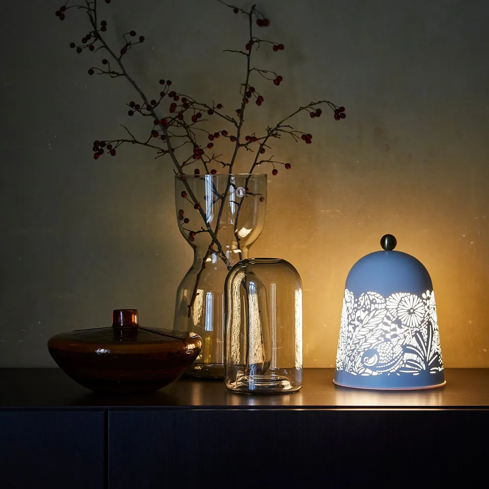 A dark shelf with a blue SOLSKUR lamp, white floral design, and transparent vase holding red-berried branches.
