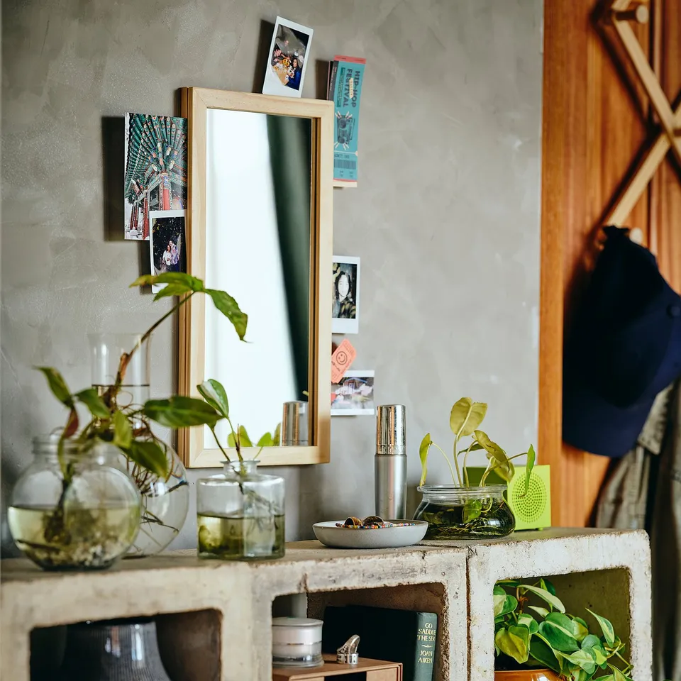 Mirror sits on table, propped by stand. It holds photos and notes in side grooves. Plants in jars surround it.