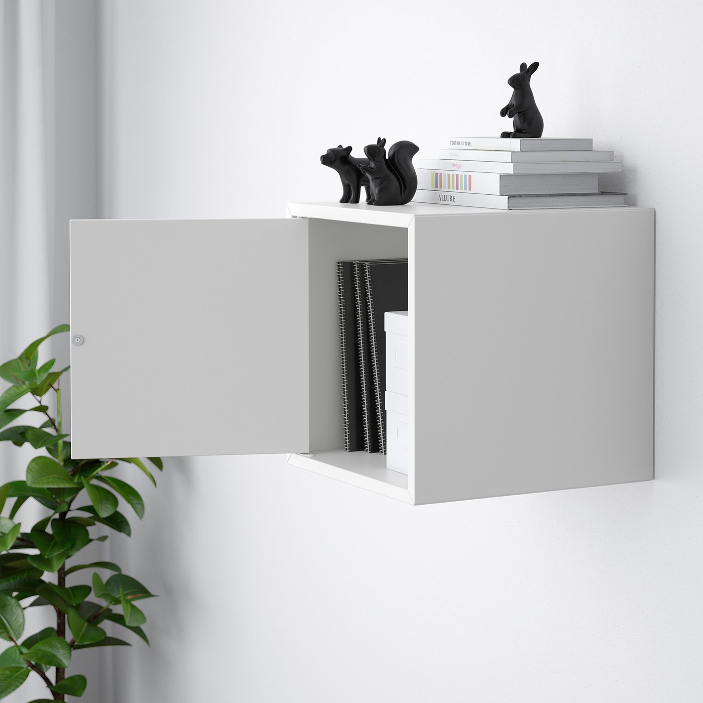 White wall-mounted cabinet with open door revealing inner storage, stacked books, and black figurines on top.