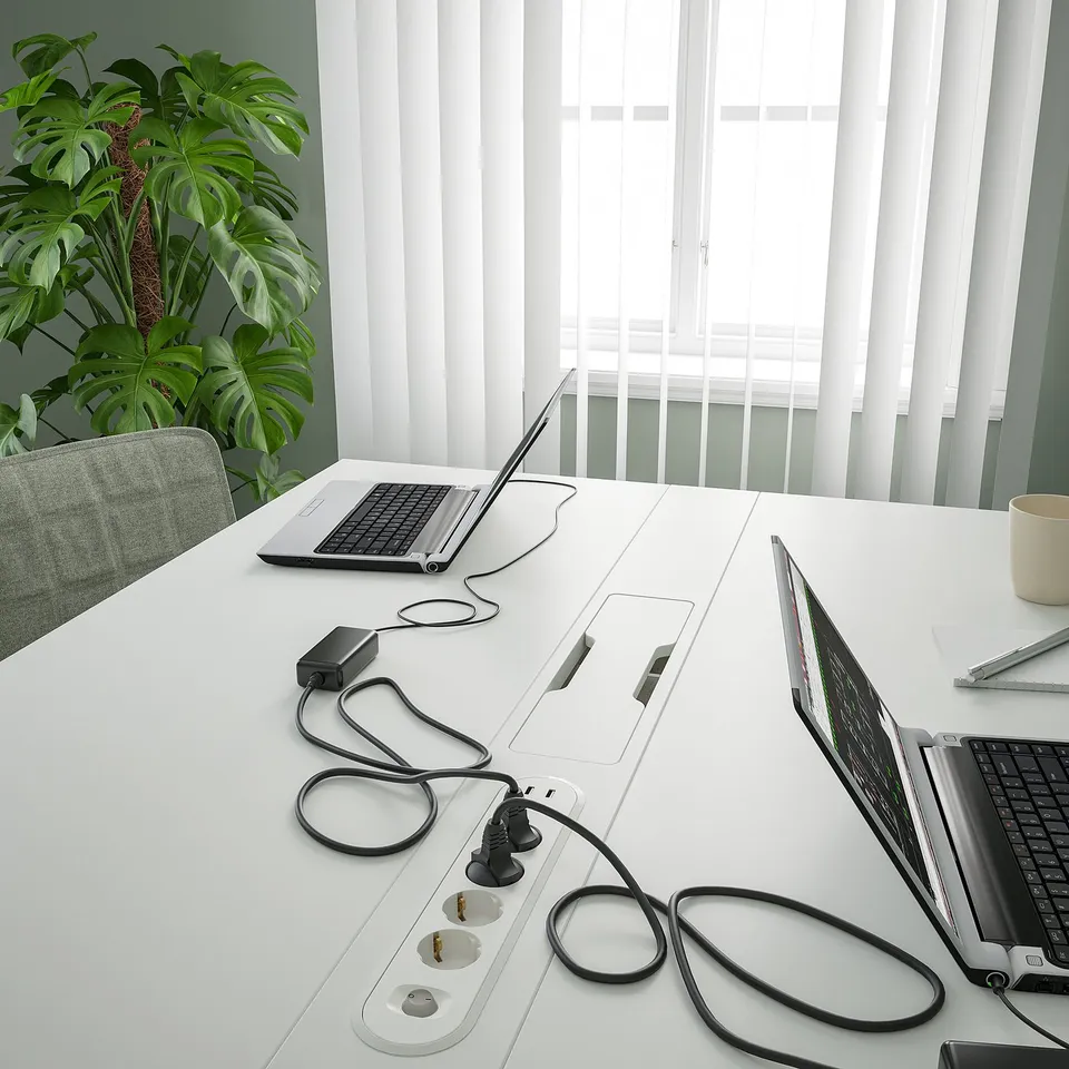 Modern conference table with built-in power outlets and cable management for laptops, positioned near a large plant and window blinds.
