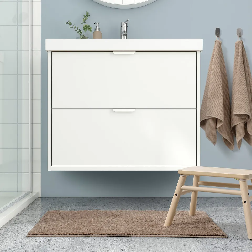 Modern bathroom with white vanity, beech stool, tan rug, & towels on hooks. Mat matches rug.