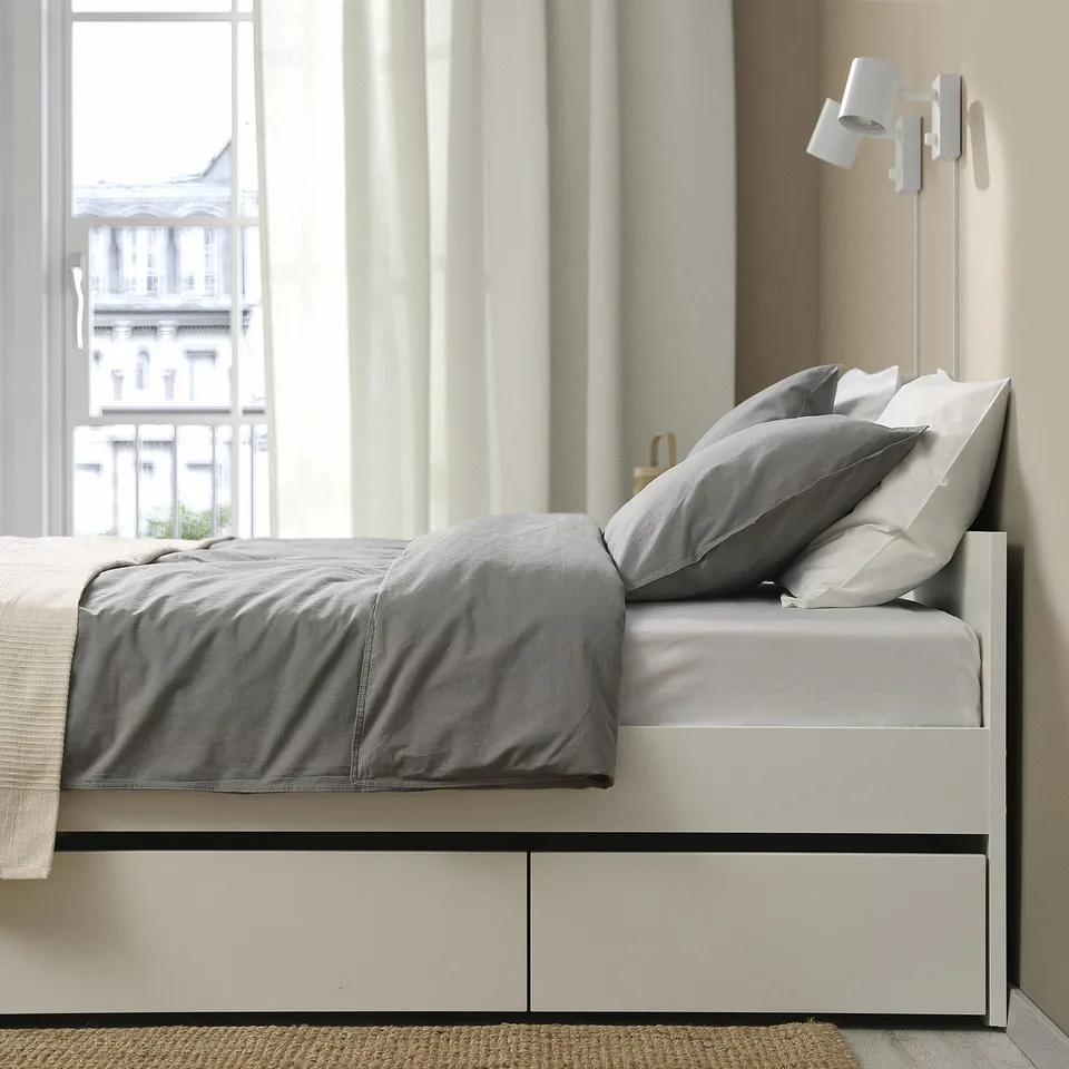 Modern bed frame with storage boxes, grey bedding, and city view from large windows.