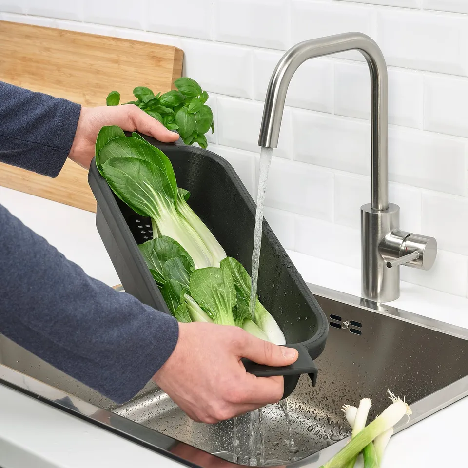 Person washes green vegetables in black LILLHAVET colander under tap over sink.