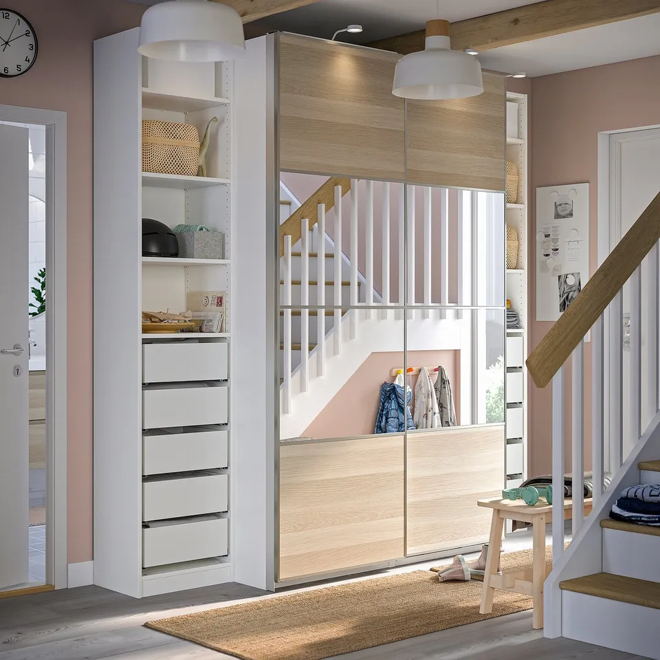 Organised hallway with white PAX wardrobe, shelves, and staircase. Drawers, baskets, and hanging space, topped with a pendant light.