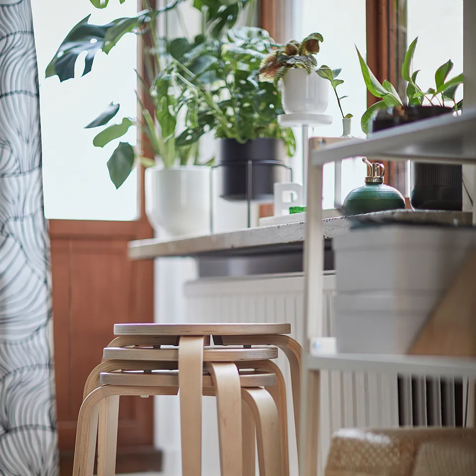Natural wooden KYRRE stools stacked near a window with plants and textiles in a cosy room.