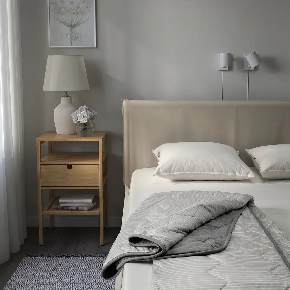 A neatly made bed with white BRANDLILJA duvet, grey accents, wooden bedside table holding a large lamp with a beige shade, and a vase with flowers.
