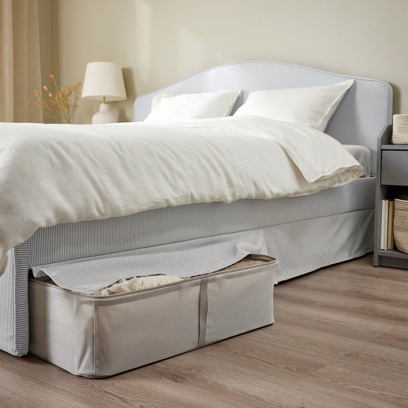 A cosy bed with a curved headboard, white sheets, and a striped bed skirt covering storage underneath. A lamp with flowers sits on the bedside table.
