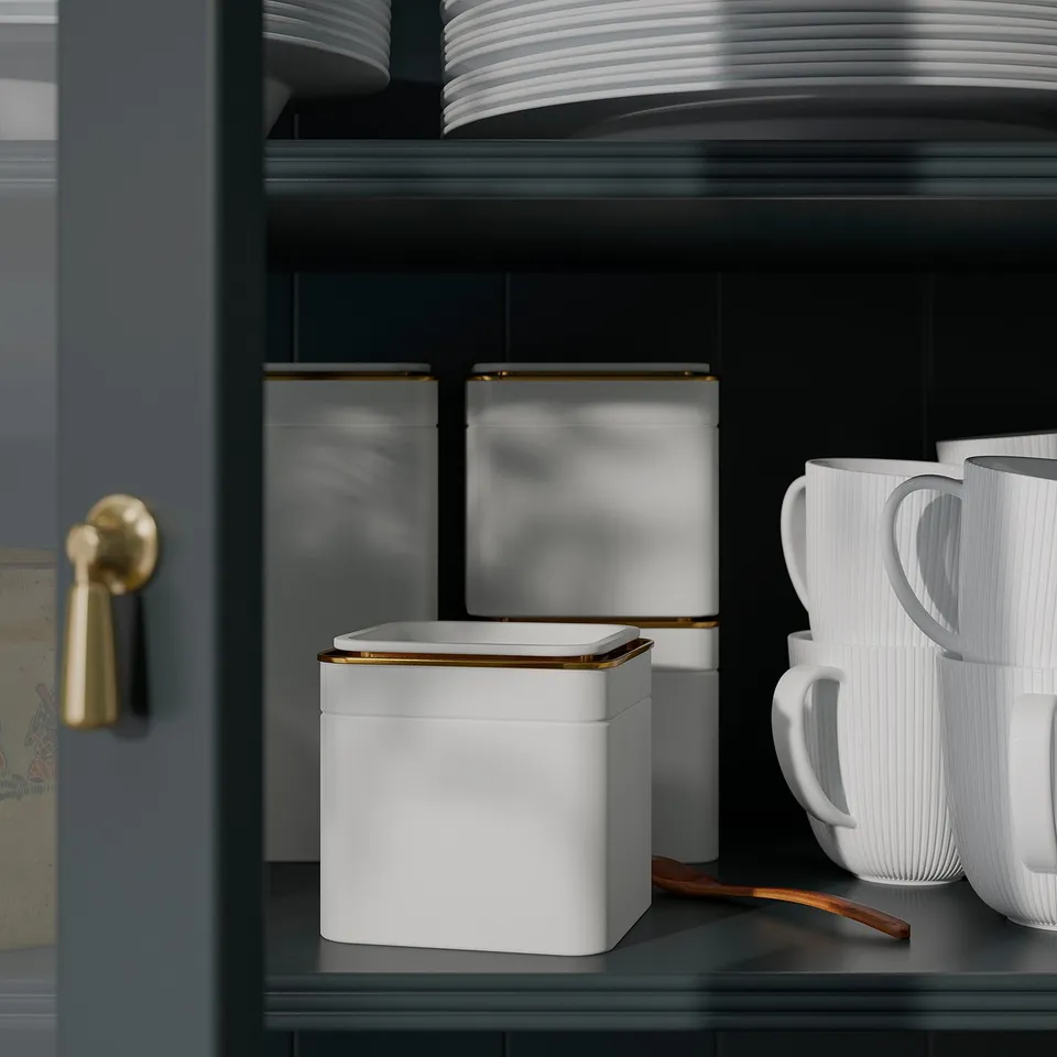 Kitchen shelf with three BLOMNING tins, white with gold trim, storing dry goods, next to stack of white mugs.