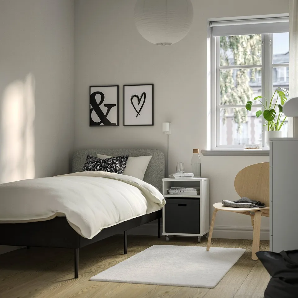 Grey bed with white sheets, wooden chair, bedside table with books & glass, window with plant. Simple, well-lit room.