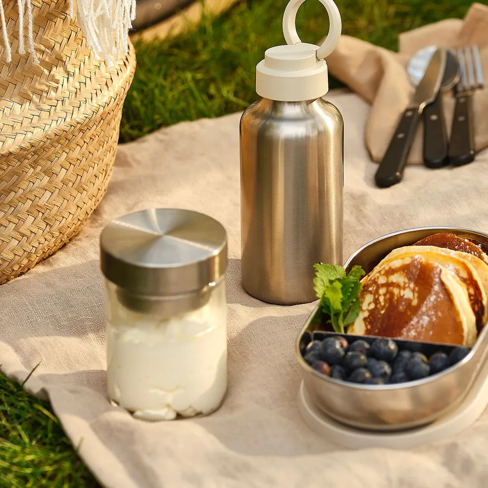 Picnic setup with stainless steel water bottle, yoghurt jar, and berry dish on cloth, wicker basket nearby.