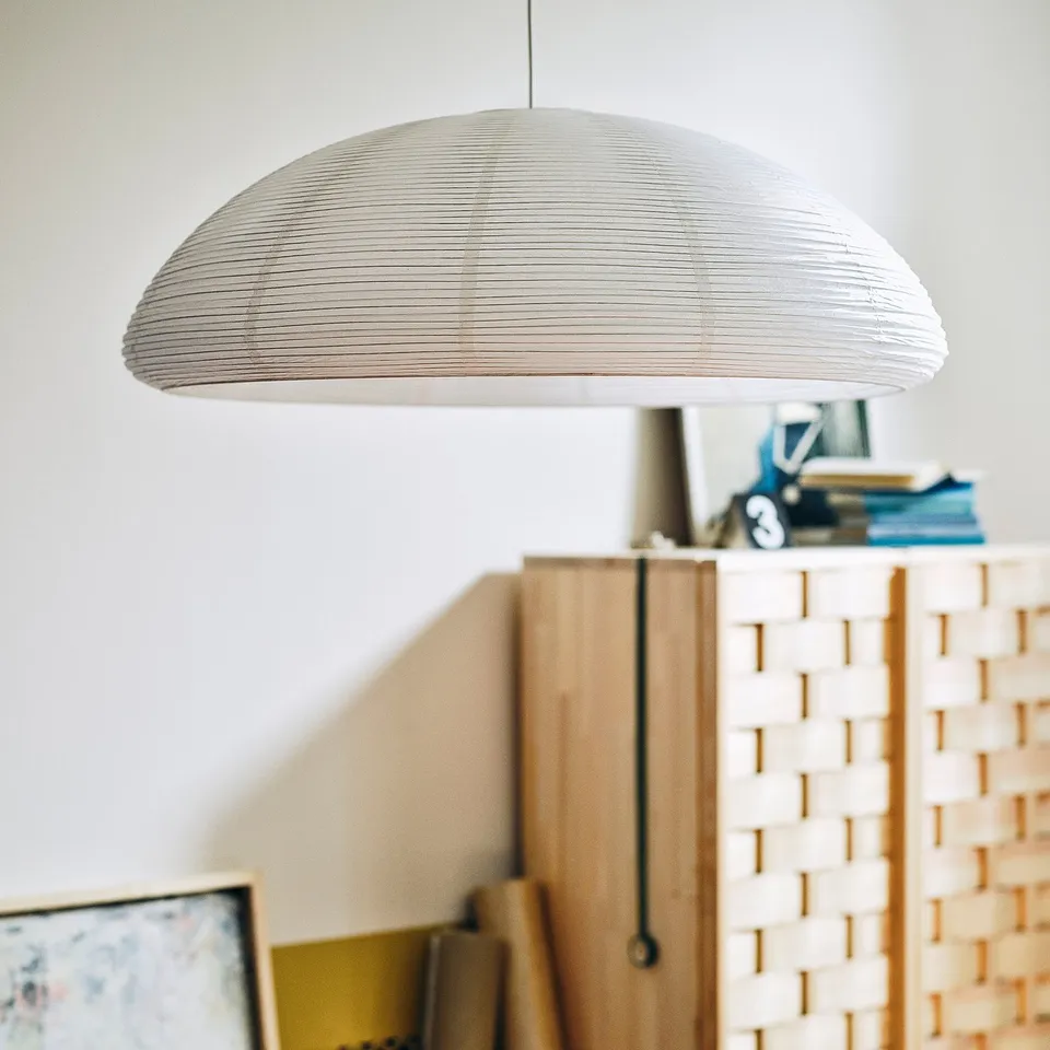 IKEA PS 2026 half-moon rice paper lamp hangs large, casting soft light. Below, wooden room divider with woven design and stacked books.