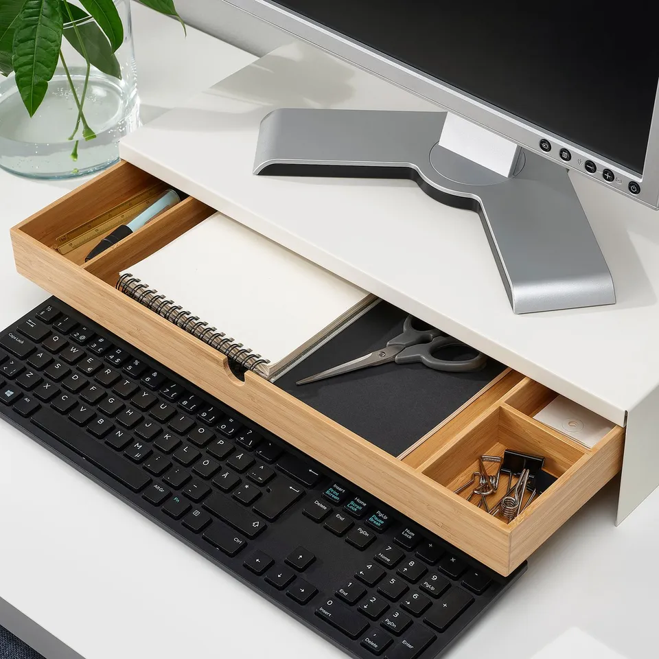 Desk with open storage drawer under monitor stand holding stationery, scissors, and notepads. Keyboard sits below, keeping workspace tidy.